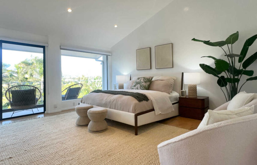 Vaulted primary bedroom suite staging in Malibu with neutral platform bed, bouclé ottomans, plush lounge chairs, and balcony access.