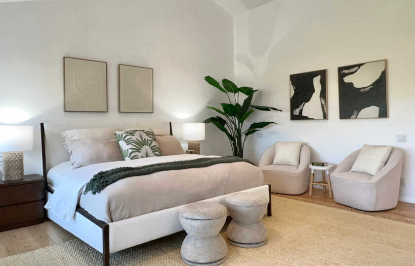 Staged primary bedroom suite showcasing neutral platform bed, woven nightstands, bouclé accent chairs, and botanical wall prints.