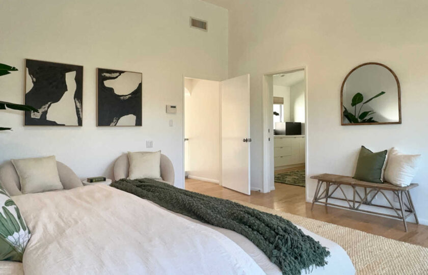 Malibu primary suite staging showing woven entry bench, arched wall mirror, abstract black-and-white art, and green accent throw.
