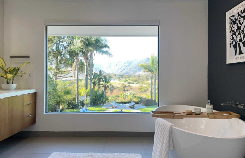 Luxury primary bathroom staging in Malibu showcasing freestanding soaking tub, wooden bath caddy, and floor-to-ceiling mountain window view.