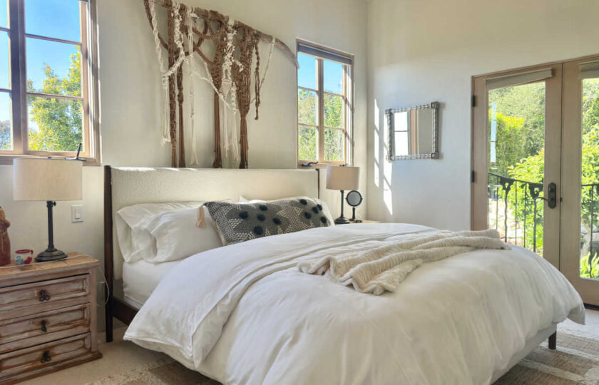 Sunlit Spanish Colonial staged bedroom with a white upholstered bed, macramé wall hanging, woven textures, and balcony access.