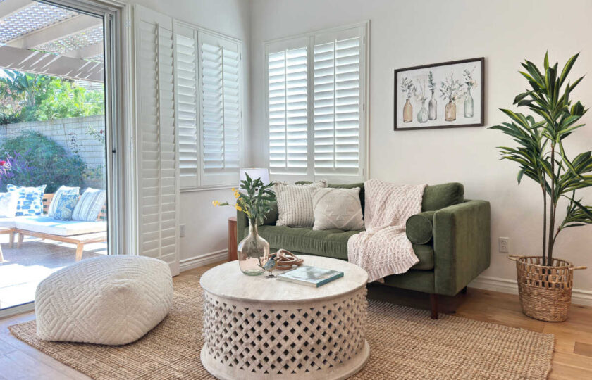 Cozy family room staged with an olive green velvet sofa, carved wood coffee table, woven jute rug, and plantation shutters.