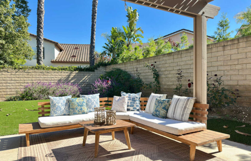 Backyard outdoor living space staged with a wooden sectional sofa, patterned throw pillows, and an outdoor rug under a shade pergola in Dos Vientos.