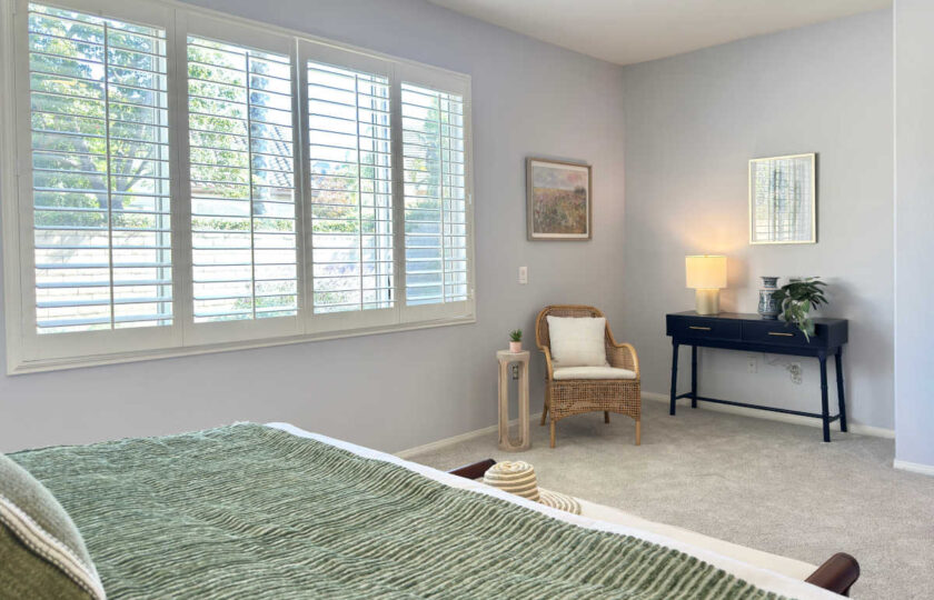 Dos Vientos Home Staging primary suite featuring large plantation shutters, a rattan accent chair, and a black console desk.
