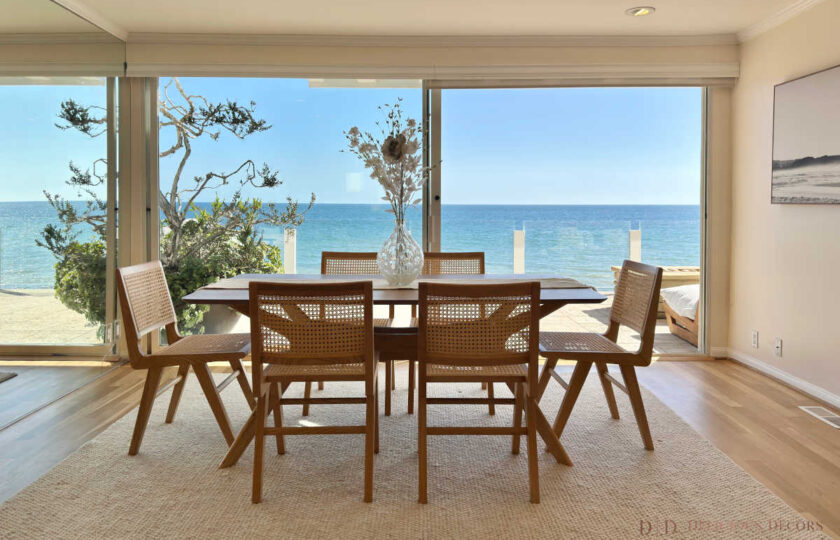 Oceanfront dining area staged with woven cane chairs, a wood table, neutral area rug, and floor-to-ceiling glass sliding doors overlooking Malibu waters.