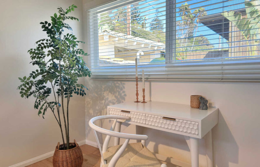 Guest bedroom desk nook staged with a white textured writing desk, woven wishbone chair, and a tall potted tree by the window.