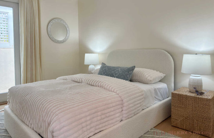 Neutral guest bedroom staged with a curved cream boucle bed frame, ribbed white comforter, and woven bedside tables.
