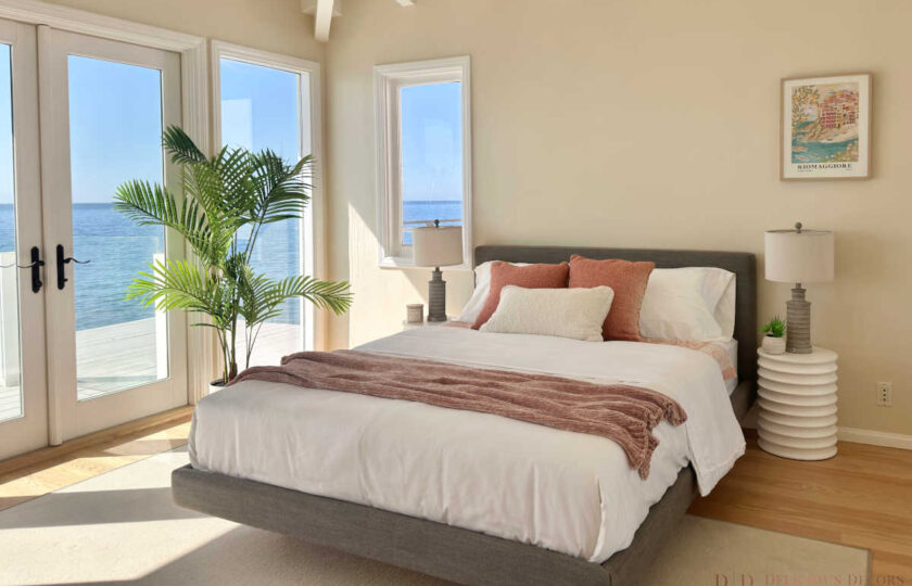 Guest bedroom staged with a charcoal grey upholstered bed frame, coral throw blanket, lush palm plant, and double doors opening to the Malibu ocean deck.