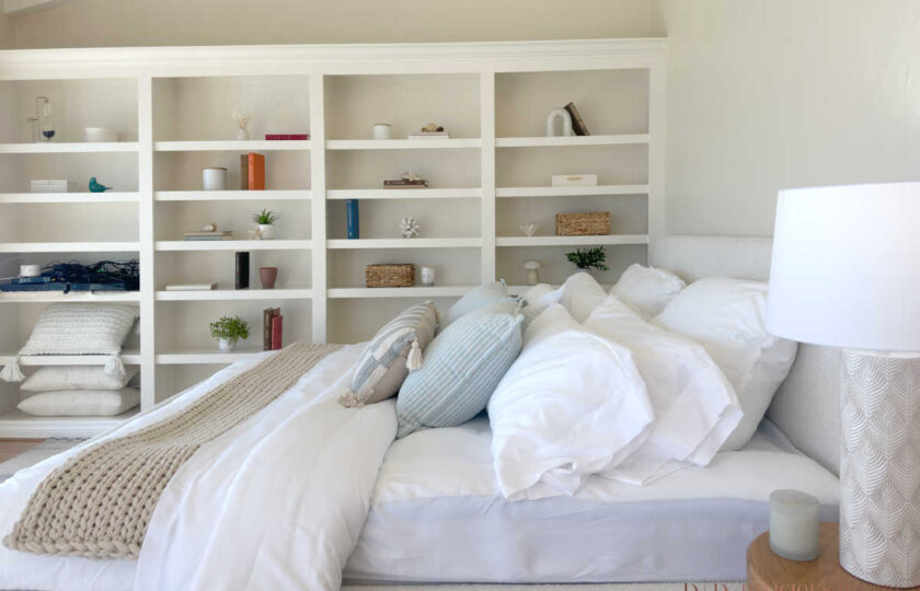 Close-up bedroom staging detail showing plush white bedding, soft blue decorative pillows, custom built-in bookshelves, and a textured nightstand lamp.