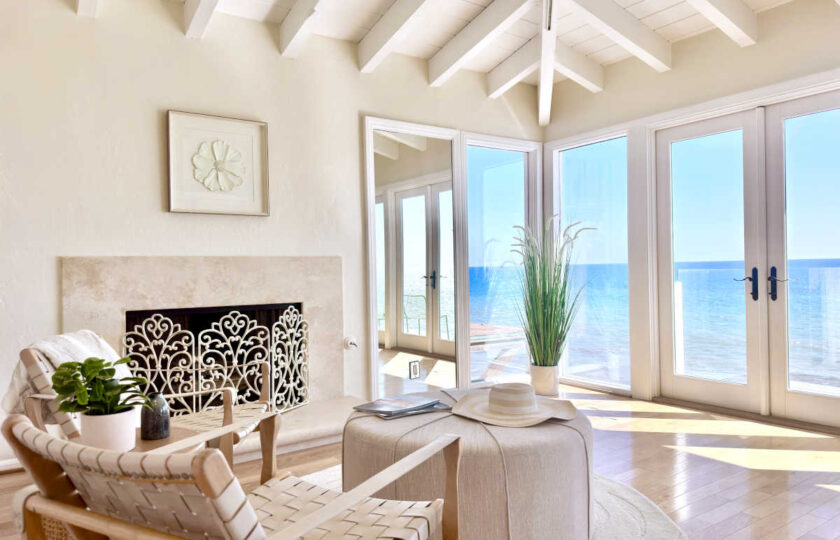 Oceanfront primary suite sitting room staged with woven leather accent chairs, a round upholstered ottoman, stone fireplace, and balcony doors.