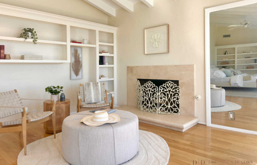 Light-filled primary suite seating nook featuring a decorative wrought iron fireplace screen, round ottoman, light wood flooring, and white beam ceilings.