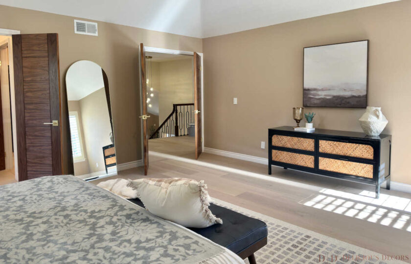 Spacious primary bedroom staging showcasing an arched full-length floor mirror, double entry doors, and a woven storage credenza.