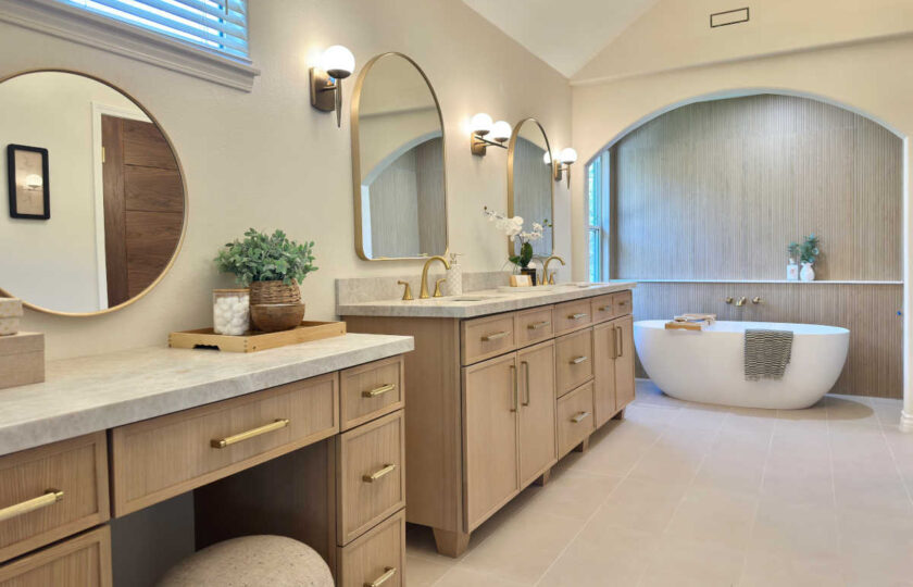 Luxury primary bathroom staged with light oak double vanities, brass arched mirrors, vaulted ceilings, and a freestanding soaking tub in Westlake Village.